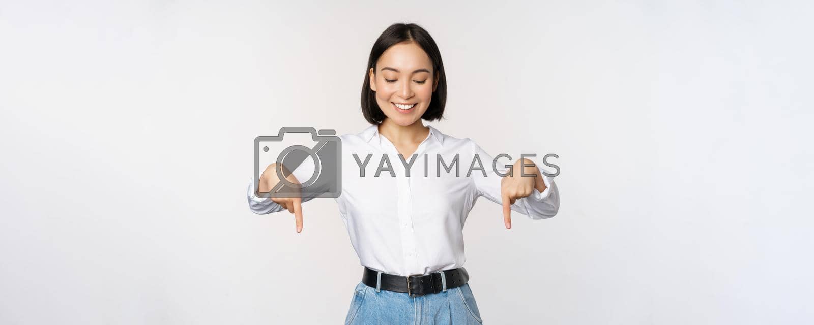Royalty free image of Portrait of happy asian woman pointing fingers down and looking below at advertisement, showing info banner, advertising, standing over white background by Benzoix