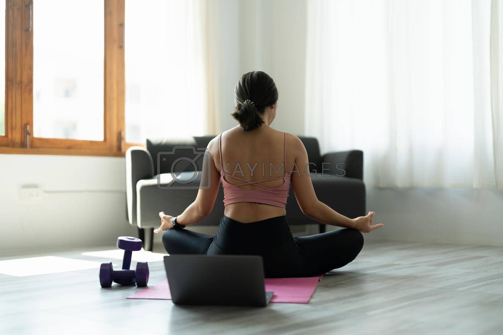 young woman in sportswear meditating while sitting in lotus pose on yoga mat at home.