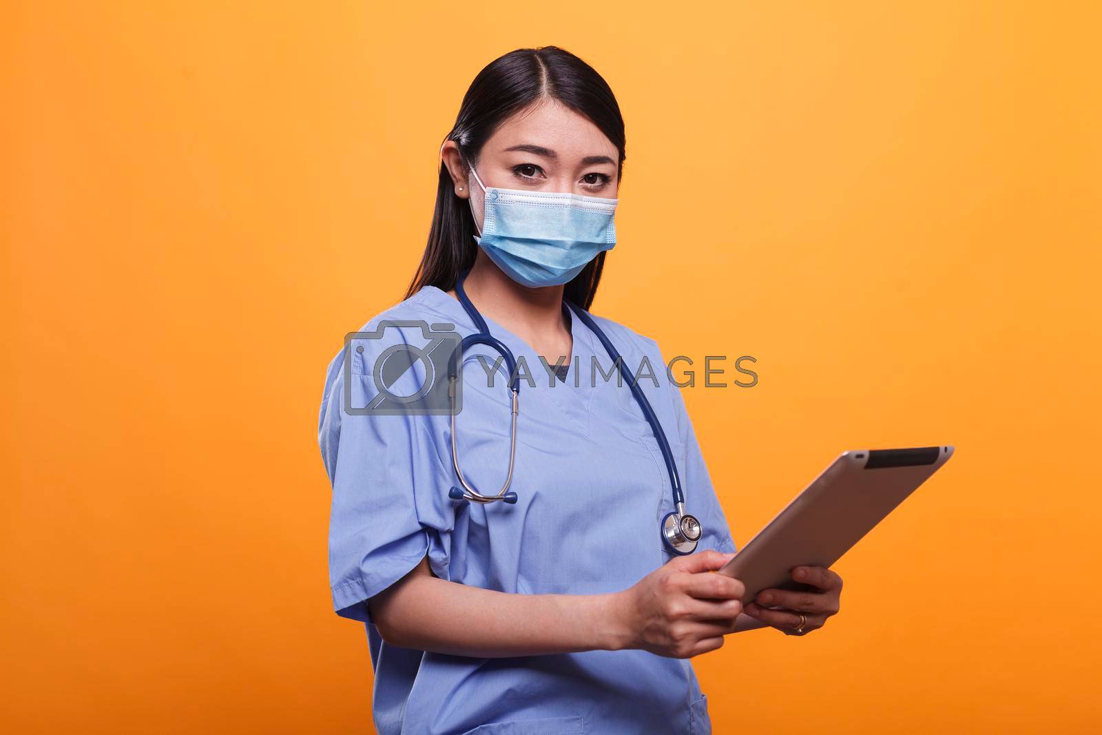 Healthcare clinic asian nurse wearing virus protection facemask and stethoscope while having modern tablet. Caretaker woman holding touchscreen pad while wearing protective mask on orange background.