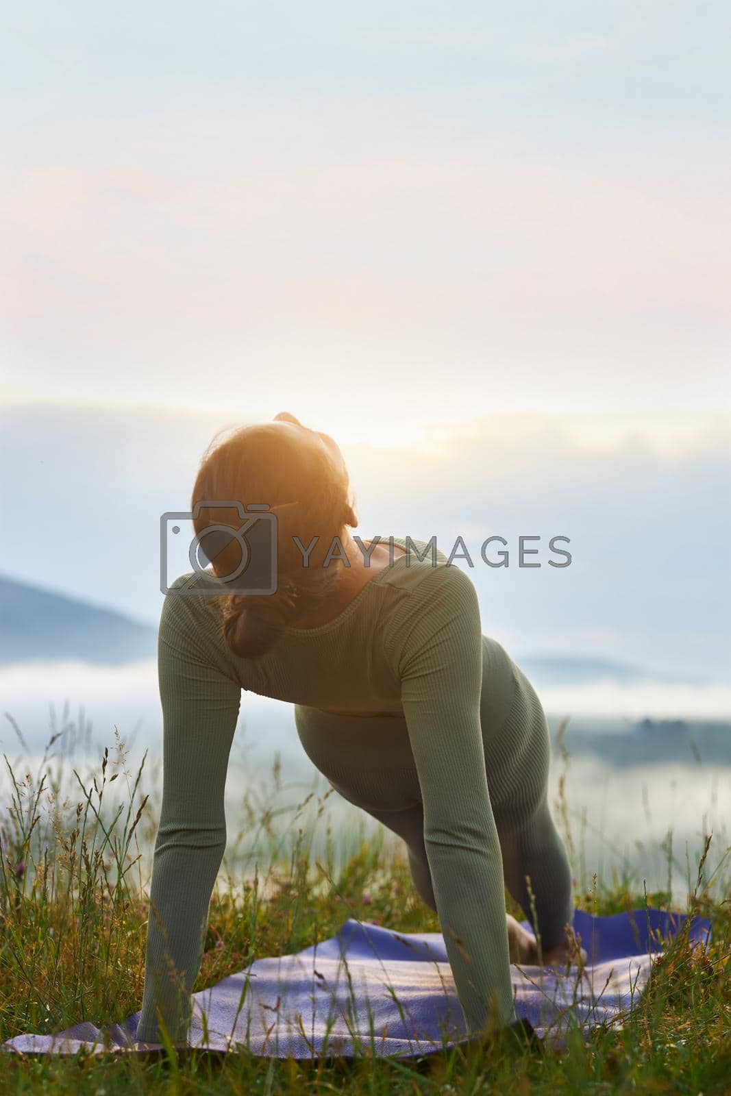 Back view of young woman practicing yoga position in mountains. Flexible and slim female standing in plank on yoga mat wearing sport suit in hills. Concept of yoga.