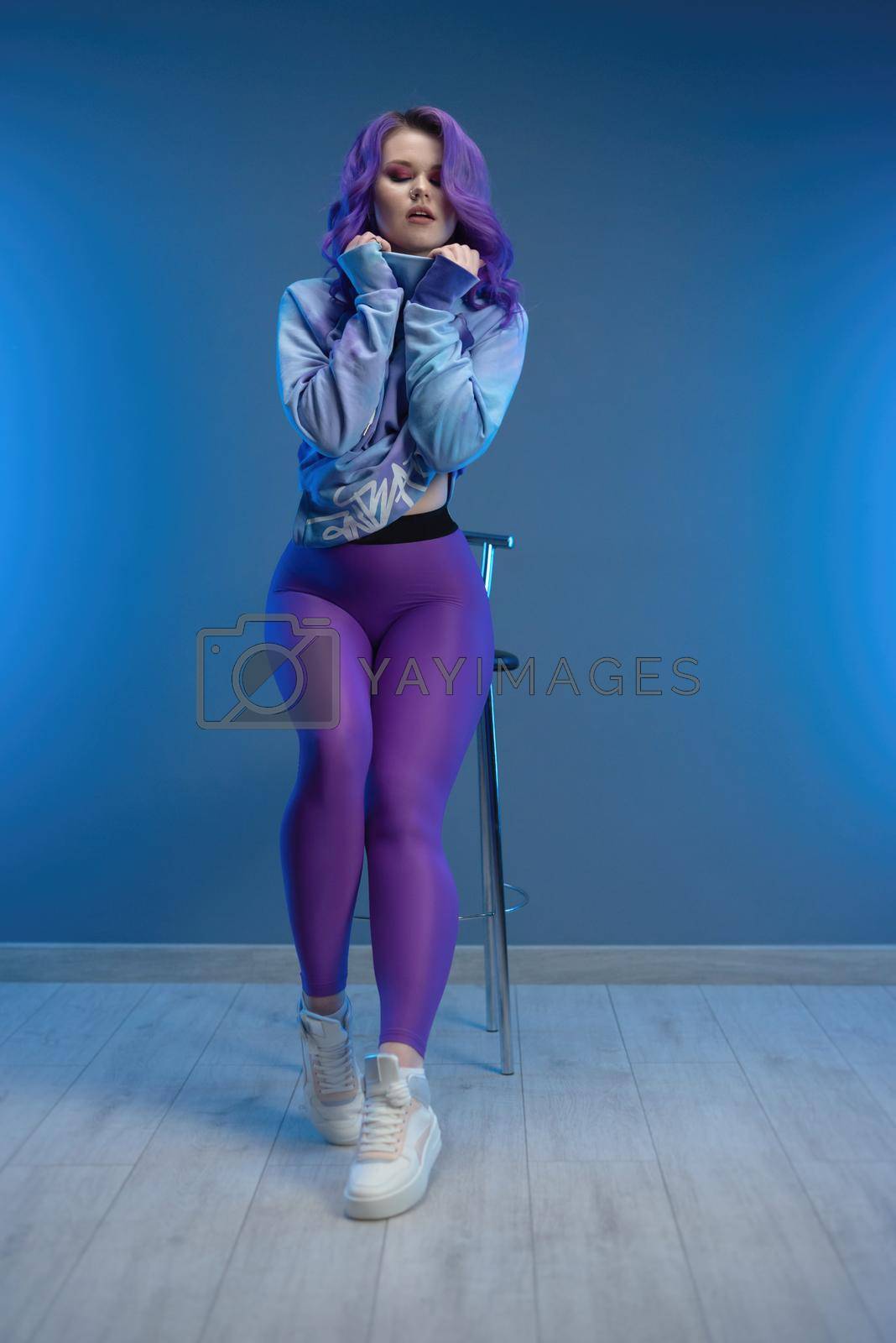 Royalty free image of a girl in stylish purple sportswear and with purple hair poses sexually on a bar stool by Rotozey