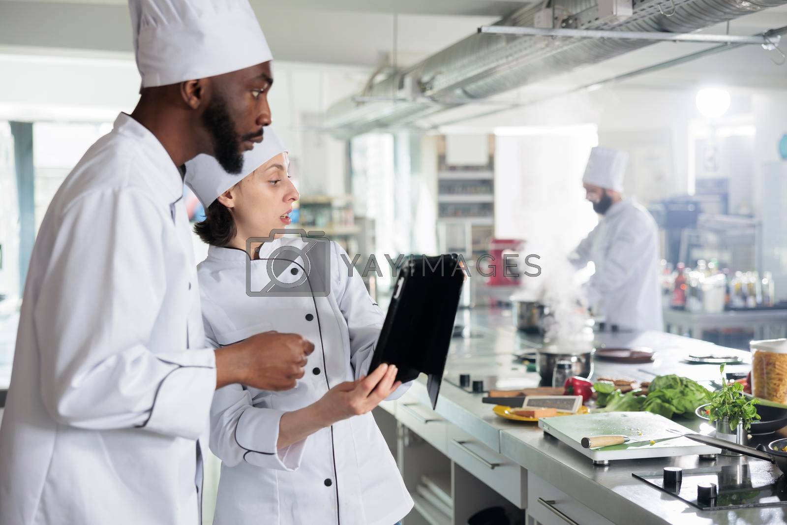 Gourmet cuisine experts with modern tablet searching for dinner service dish ideas on internet. Professional chefs with handheld touchscreen device preparing ingredients for evening dinner service.