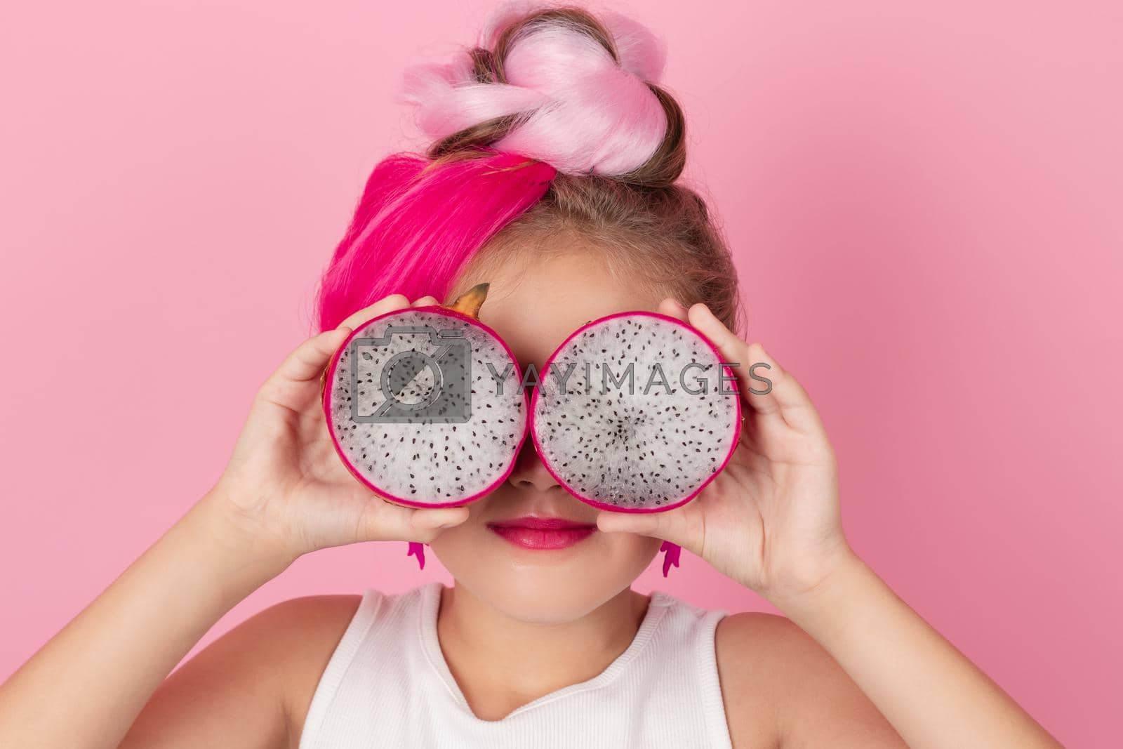 Royalty free image of Close-up portrait of pretty girl with pink hairstyle with dragon fruit on pink background. Studio shot of charming tween girl with pink make up enjoying juicy red pitaya. exotic Pitahaya fruit by oliavesna