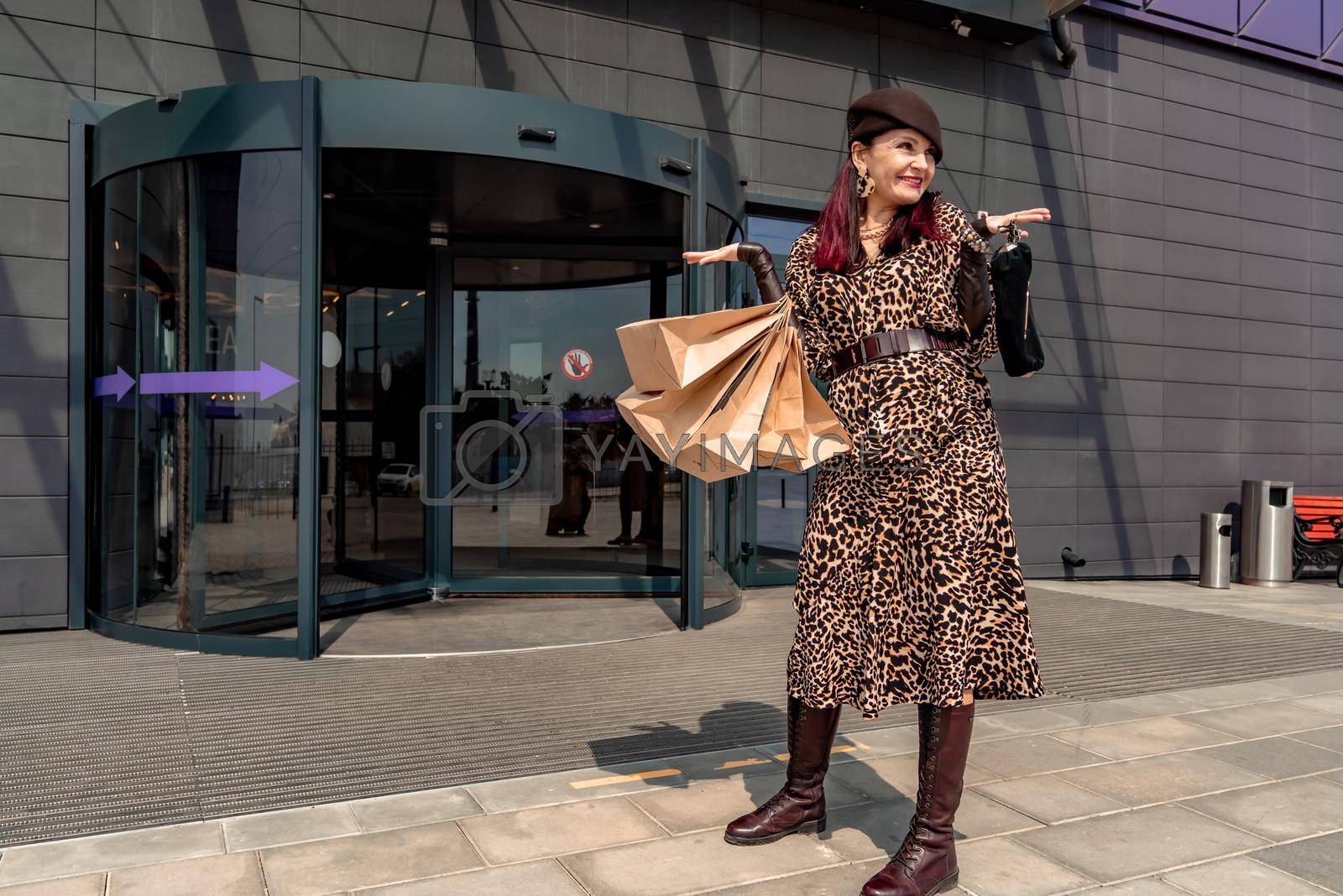 Royalty free image of A happy shopaholic girl keeps her bags near the shopping center. A woman near the store is happy with her purchases, holding bags. Dressed in a leopard print dress. Consumer concept. by Matiunina