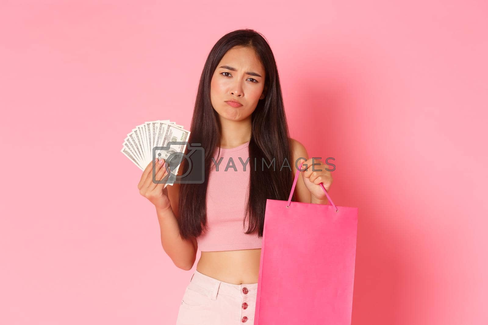 Royalty free image of Beauty, fashion and lifestyle concept. Portrait of disappointed, sulking asian girl with money and shopping bag looking unamused, tried to cheer up with buying new clothes, pink background by Benzoix