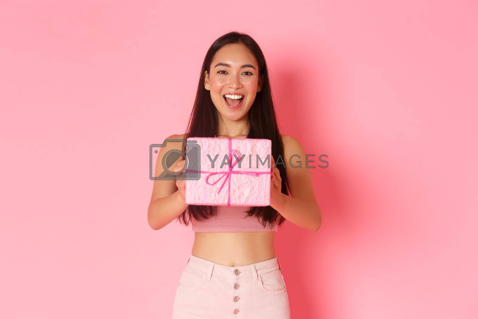 Holidays, celebration and lifestyle concept. Waist-up of cheerful beautiful asian girl making gift, giving friend birthday present in wrapped box, standing pink background, congratulate.