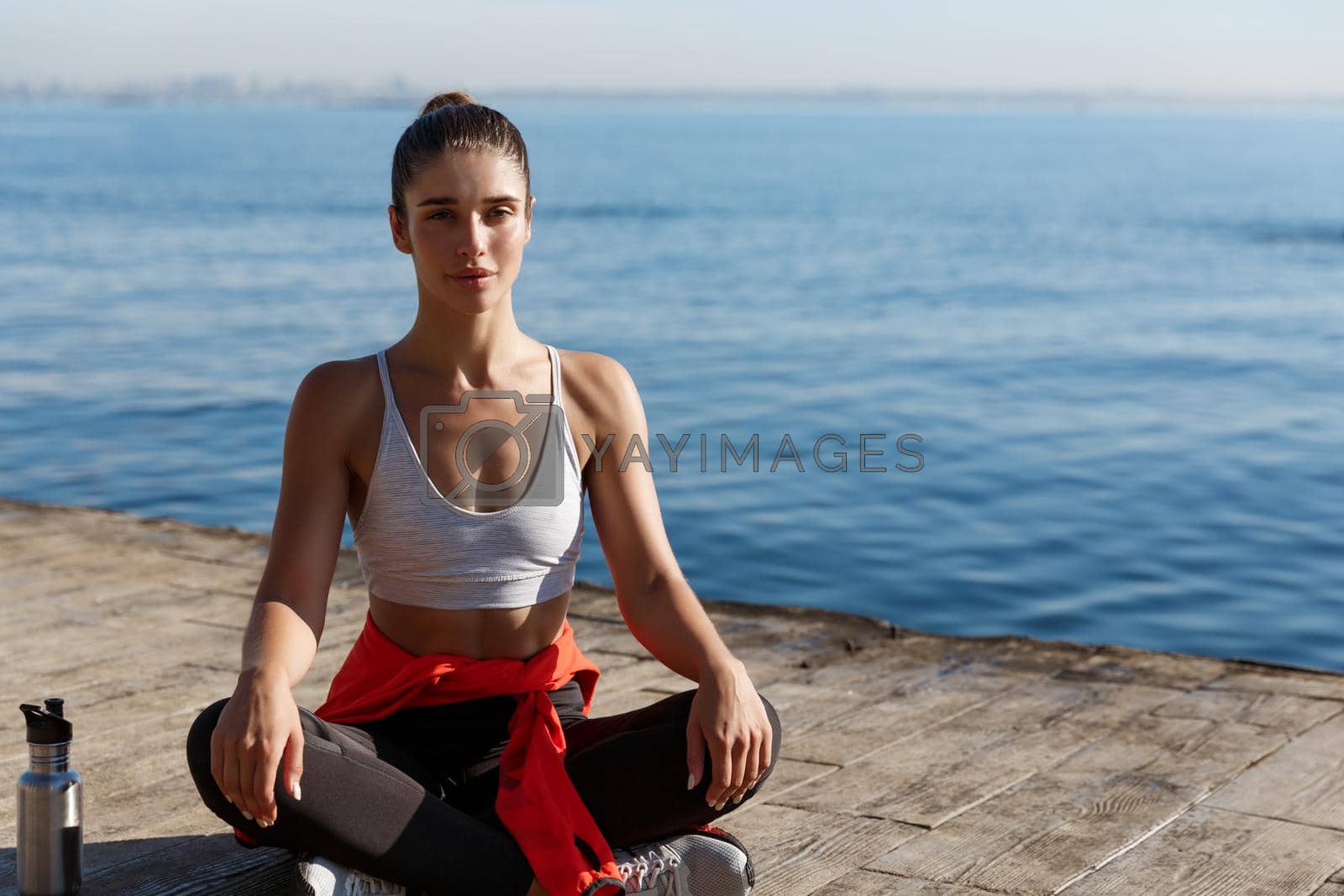 Outdoor shot of relaxed young woman meditating near the sea, practice yoga, sitting in asana on a wooden pier floor.