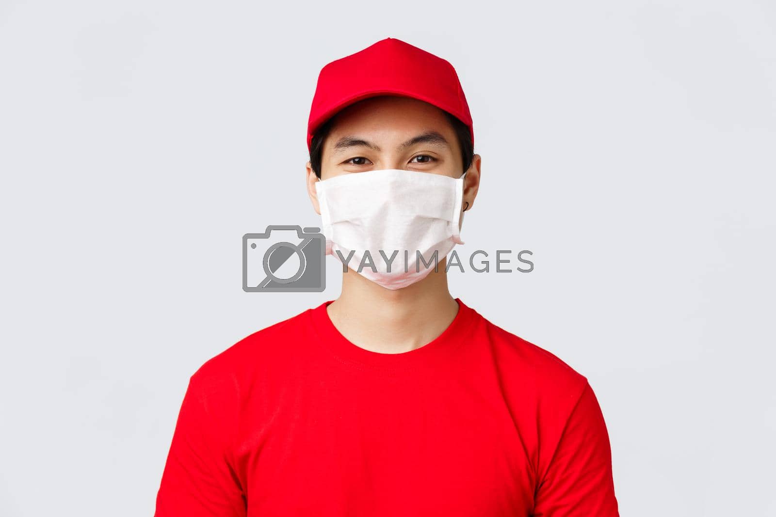 Royalty free image of Close-up portrait of cheerful asian delivery guy in medical mask and red cap uniform, t-shirt, smiling with eyes at customer, taking order using safety measures to prevent covid-19 spread by Benzoix