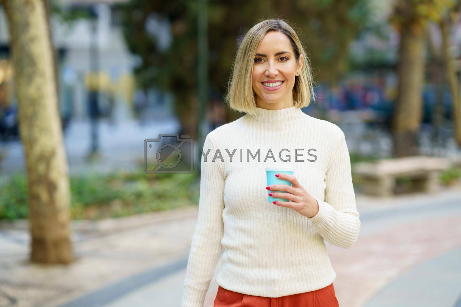 Positive female in white turtleneck with cup of takeaway beverage standing in city park and looking at camera with toothy smile