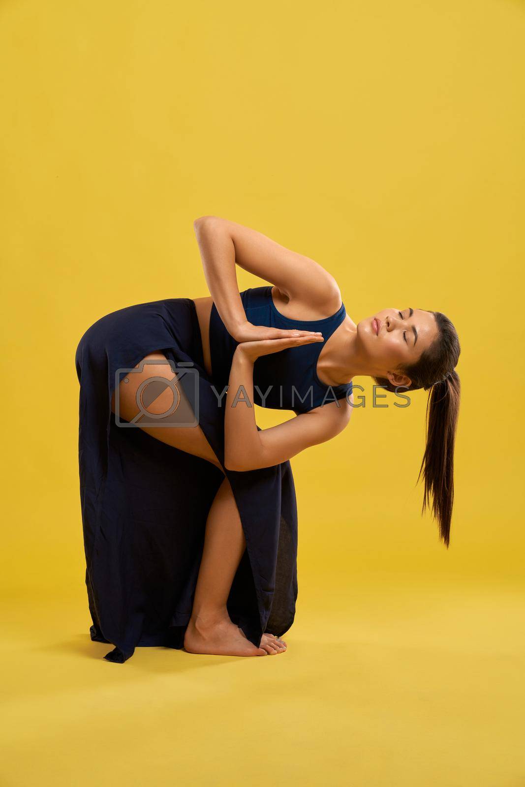 Front view of flexible girl practicing yoga pose indoors. Brunette female standing with bent legs, bending forward, with prayer hands, looking up. Concept of new age and yoga.