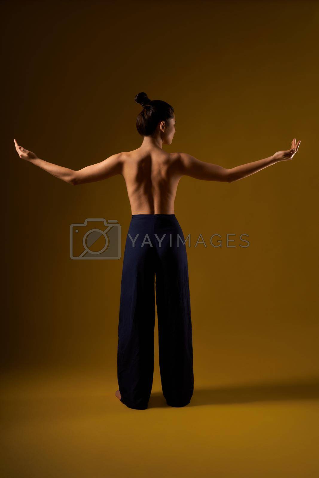Back view of slim, flexible female practicing yoga at studio. Brunette girl with bare back standing, raising arms, stretching, meditating. Isolated on yellow studio background.