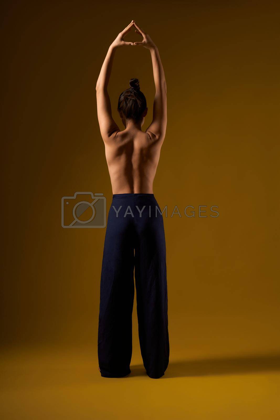 Back view of sporty female practicing yoga pose at studio. Flexible girl with bar back standing, raising hands together, meditating. Isolated on yellow studio background.