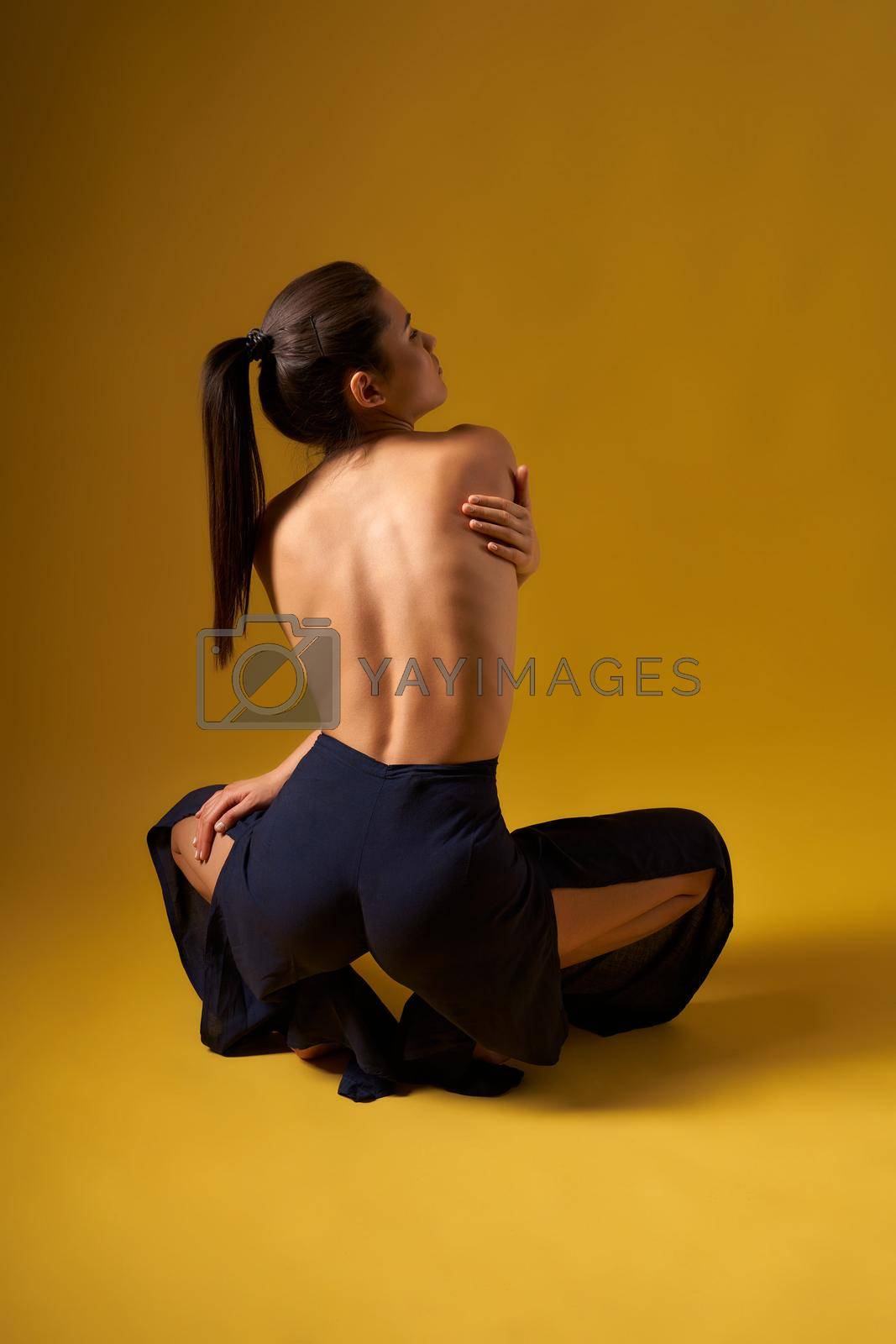 Back view of flexible girl with bare back doing yoga indoors. Brunette with pony tail sitting squant, looking forward, holding by hands. Concept of new age and yoga practicing.