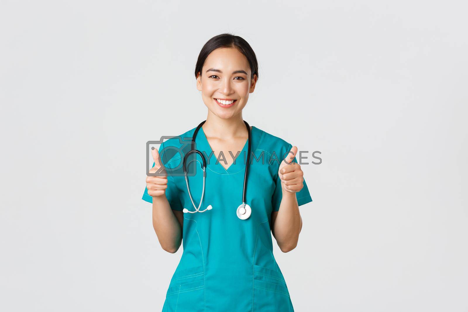 Royalty free image of Covid-19, healthcare workers, pandemic concept. Professional confident smiling doctor, female physician in scrubs showing thumbs-up with assured expression, ensure all good, perfect or excellent by Benzoix