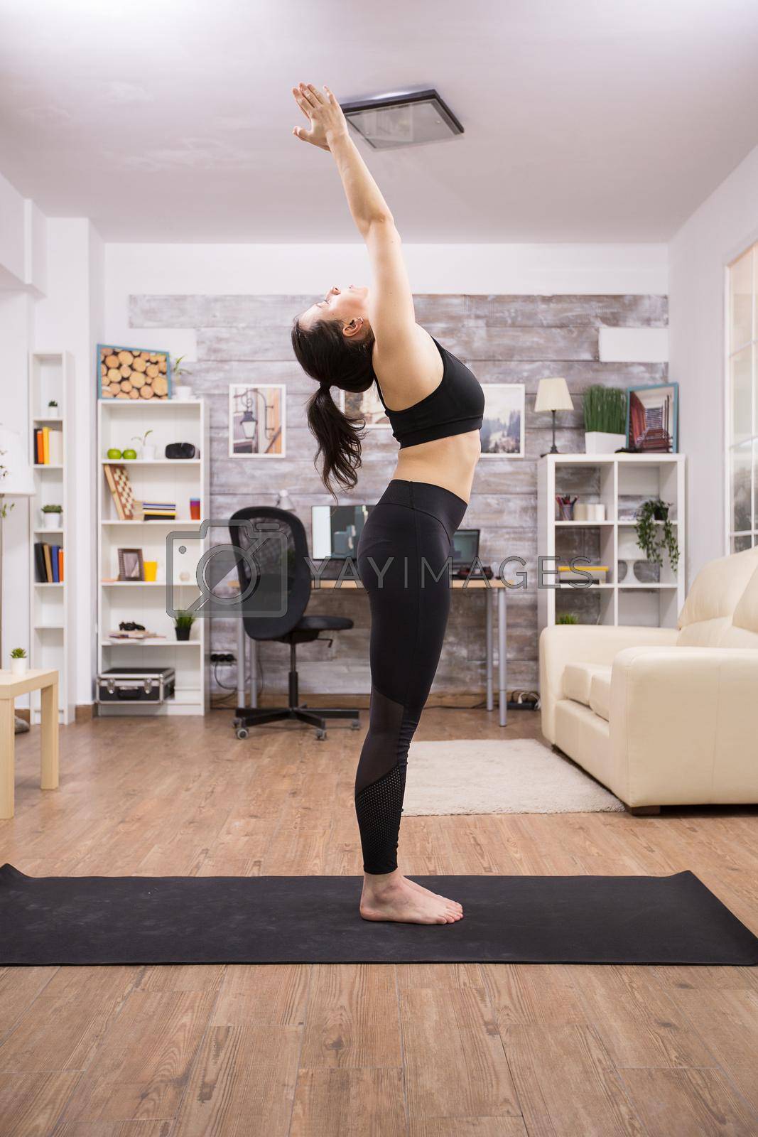Side view of young woman doing sun yoga pose in living room wearing leggings.