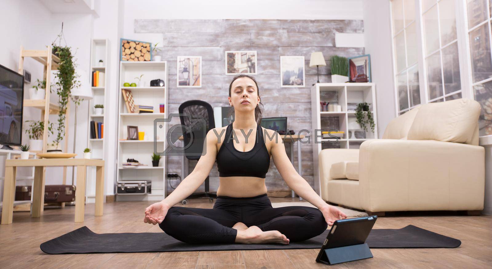 Tablet computer in front of young woman doing yoga pose sitting on mat in living room.