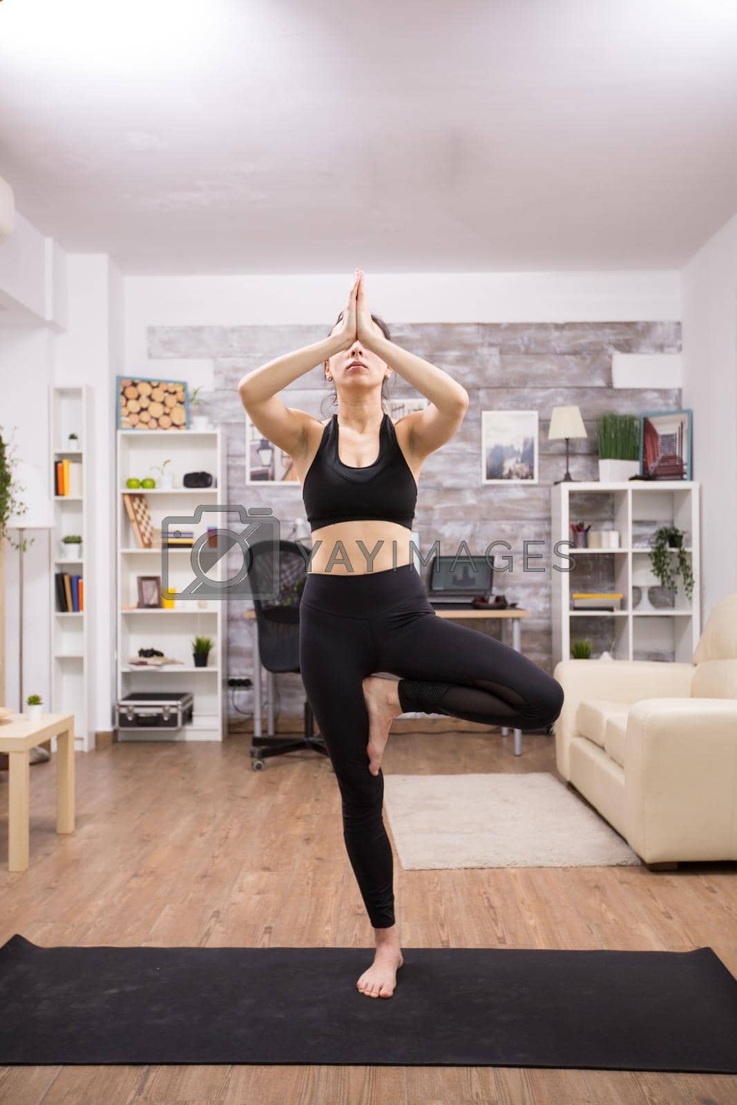 Young caucasian woman practicing tree yoga pose wearing black sportwear in home.