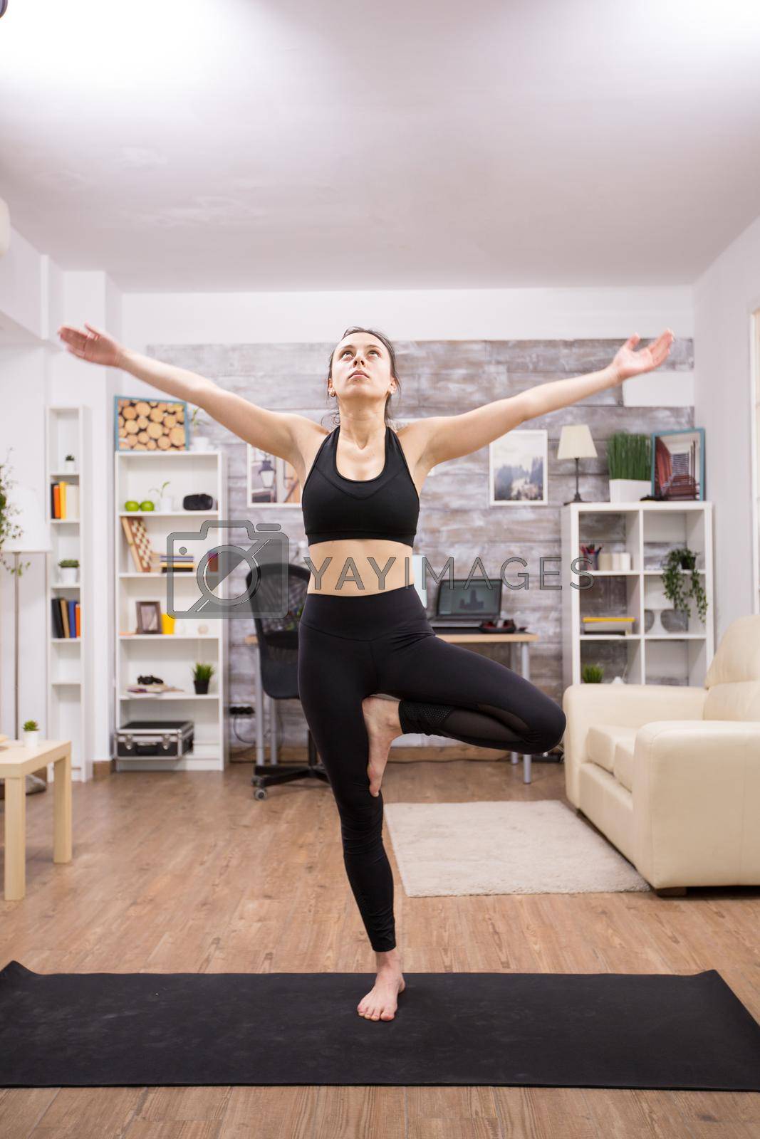 Young woman in black sportswear doing yoga tree pose in home.