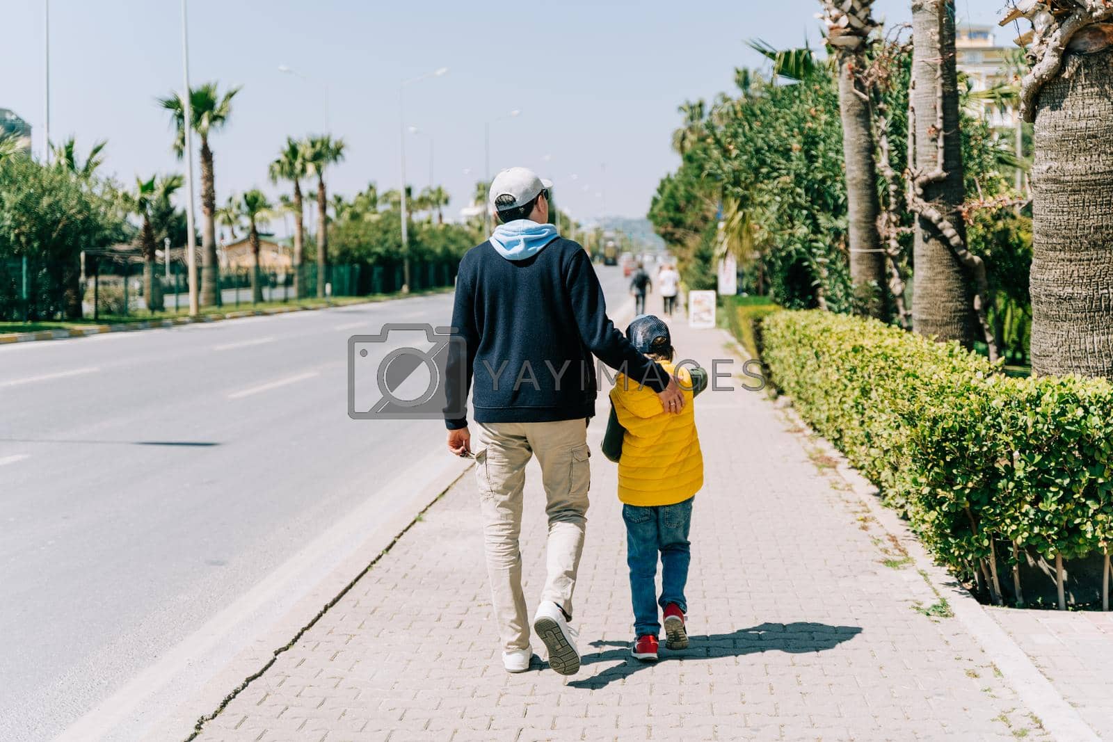 Royalty free image of Rearview of father and son walking near city road. Dad holding his kid's back when taking a stroll in the southern city. Man and boy going for promenade near highway by Ostanina