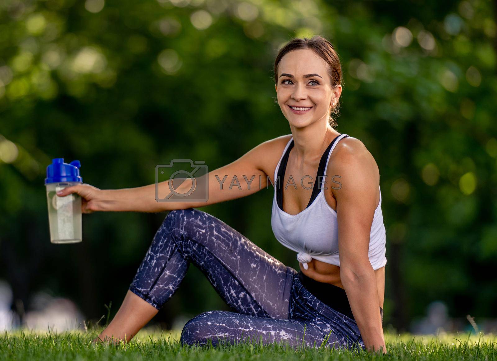 Beautiful girl sitting with water during yoga workout at the nature, looking at camera and smiling. Young woman resting after stretching fitness exercises