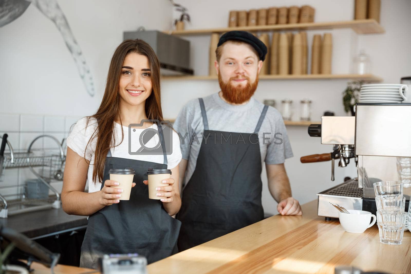 Royalty free image of Coffee Business Concept - Positive young bearded man and beautiful attractive lady barista couple giving take away cup of coffee to custome at the modern coffee shop by Benzoix