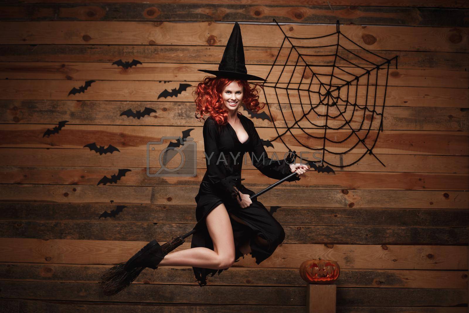 Royalty free image of Halloween witch concept - Happy Halloween red hair Witch holding magic broomstick flying gesture over old wooden studio background. by Benzoix