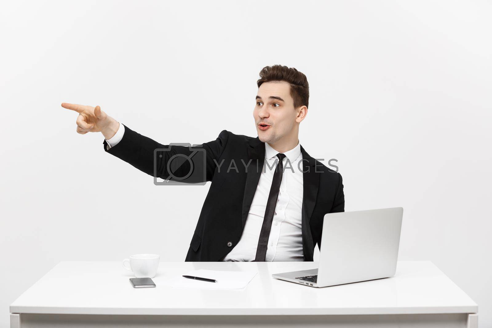 Business Concept: Portrait of handsome businessman dressed in suit sitting in office pointing finger at copy space and laptop isolated over gray background.