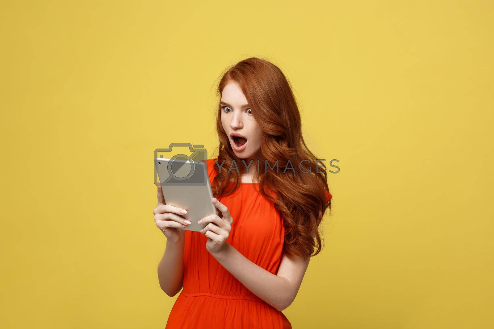 Royalty free image of Technology and Lifestyle Concept: Surprised young woman wearing orange dress clothes using tablet pc isolated on vivid yellow background by Benzoix