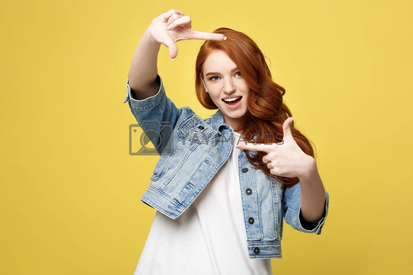 Royalty free image of Portrait of young beautiful ginger woman with freckles cheerfuly smiling making a camera frame with fingers. Isolated on yellow background. Copy space. by Benzoix
