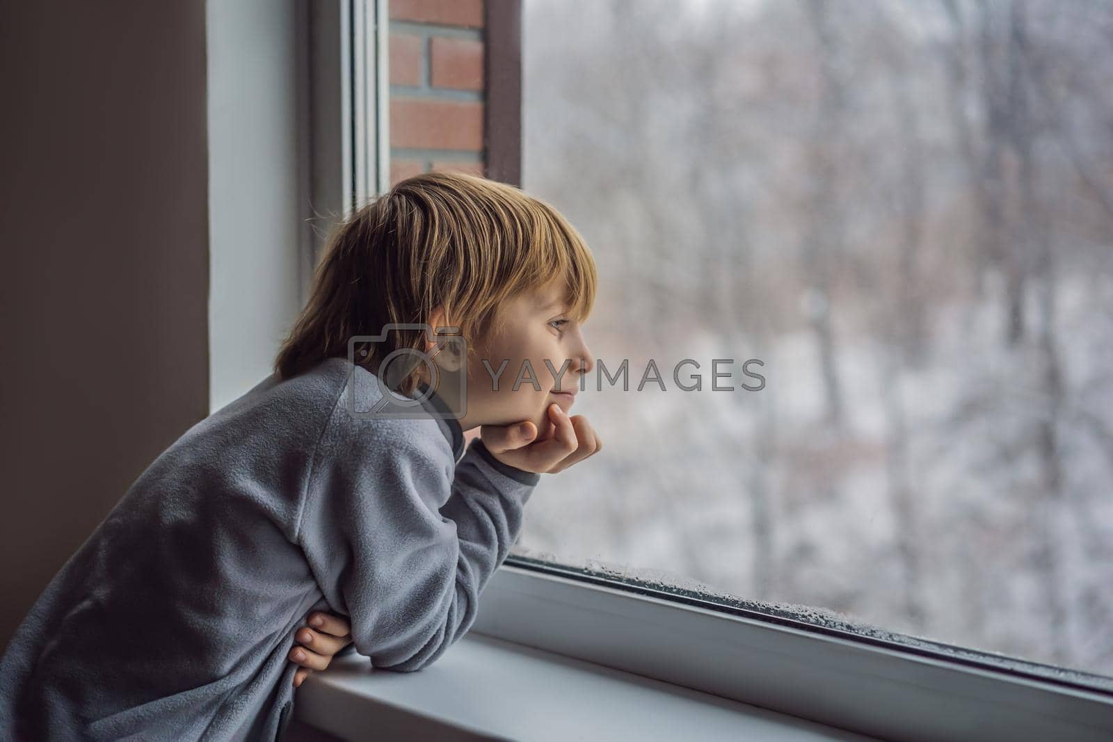 Royalty free image of Happy adorable kid boy sitting near window and looking outside on snow on Christmas day or morning. Smiling child fascinated with snowfall and big snowflakes by galitskaya