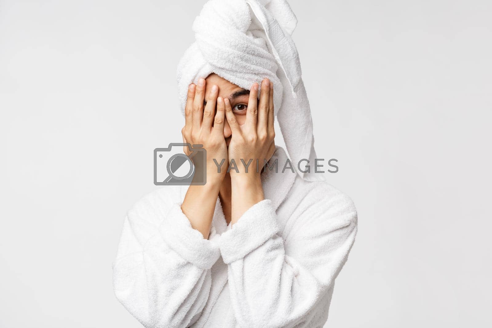 Royalty free image of Beauty, spa and leisure concept. Funny asian man in bathrobe and bath towel over head, coming from shower, hiding face behind hands and peeking with one eye curious, white background by Benzoix