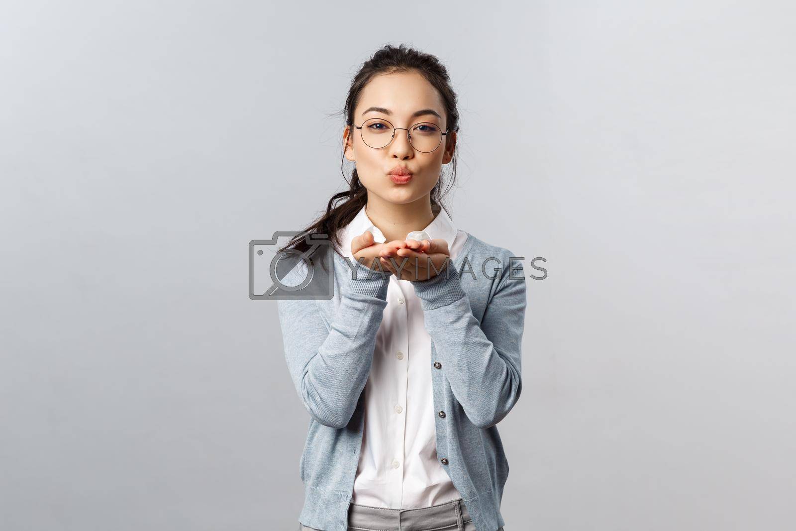 Portrait of romantic and passionate, pretty asian girlfriend in glasses send, blowing air kiss to camera with hands near folded lips, express symapthy, romance and flirt, stand grey background.