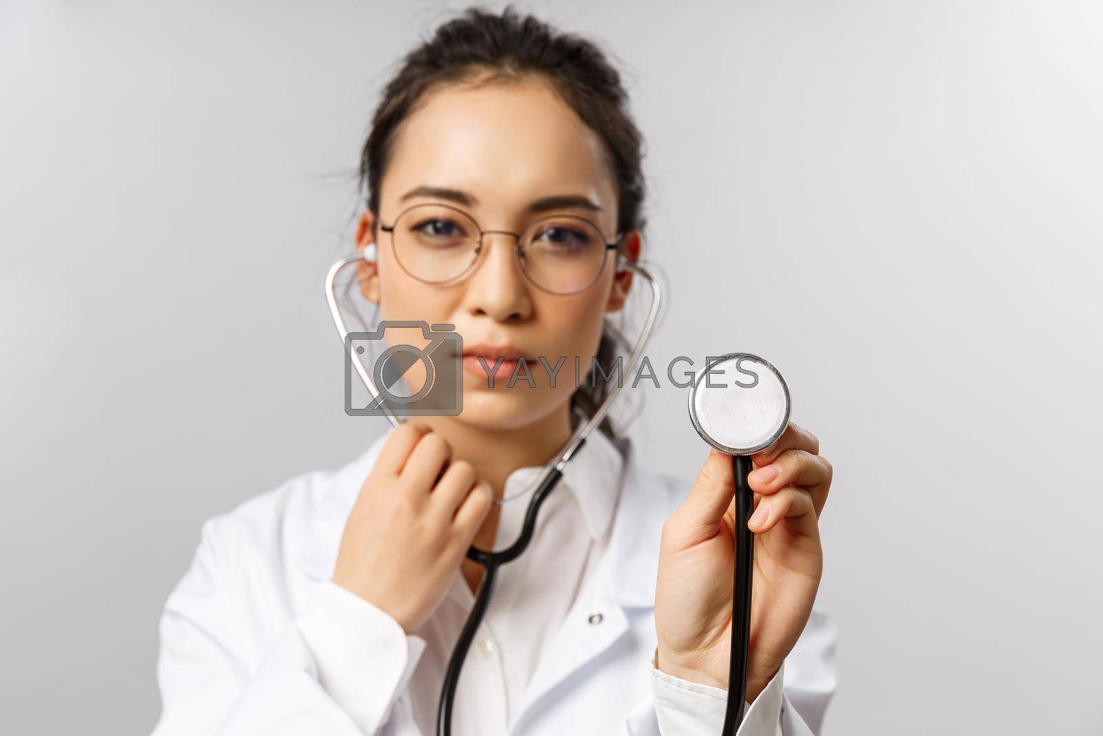 Royalty free image of Covid19, coronavirus, healthcare and doctors concept. Portrait of serious-looking female doctor during check-up of patient in ER, hospital, wearing white coat, listening to lungs with stethoscope by Benzoix