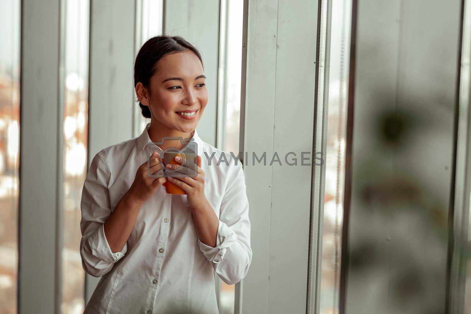 Royalty free image of Smiling asian business woman drinking coffee standing near window at office by Yaroslav_astakhov