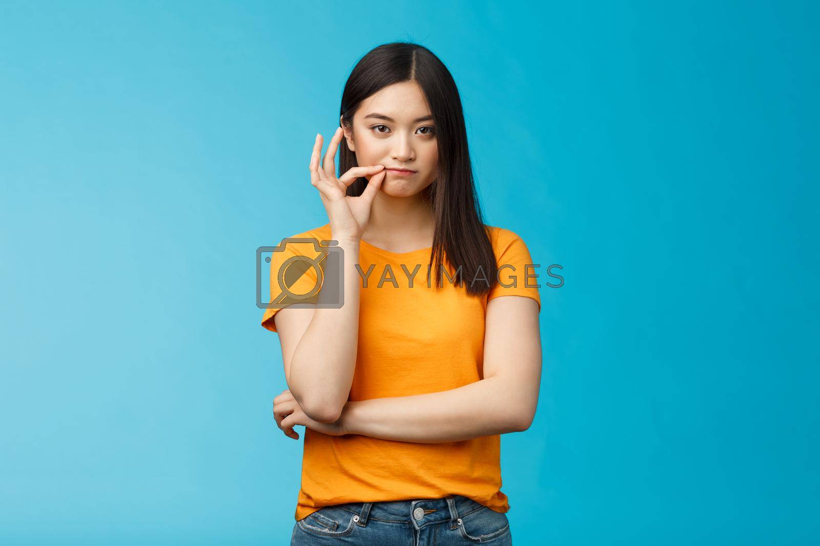 Royalty free image of Serious-looking asian female friend promise keep secret, seal lips, hold zip near mouth look focused determined, stay silent and speechless, stand wearing yellow t-shirt blue background by Benzoix