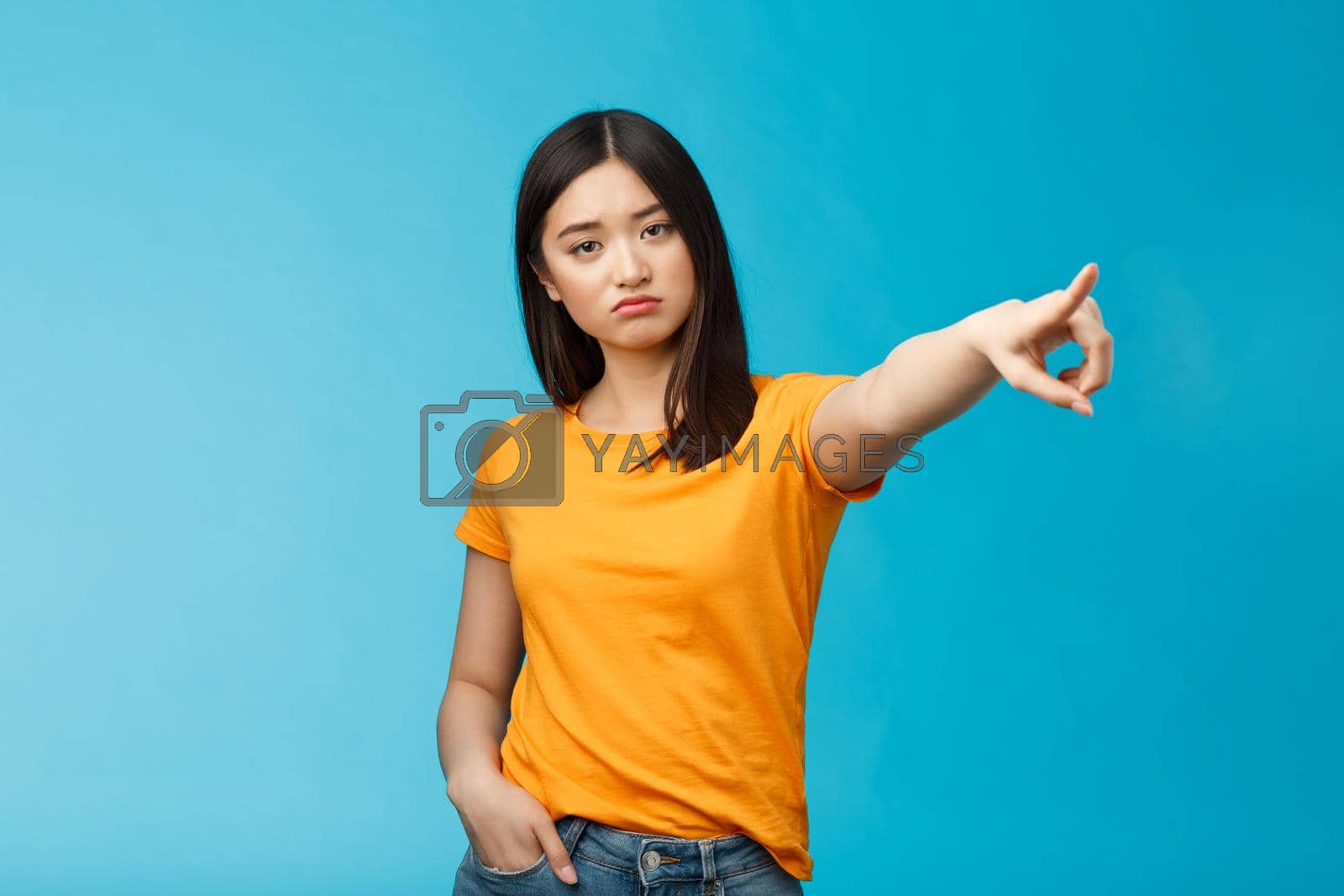 Royalty free image of Go away from here. Serious-looking arrogant displeased bossy asian girl showing way-out, pointing sideways index finger look camera demand leave, stand blue background ignorant by Benzoix
