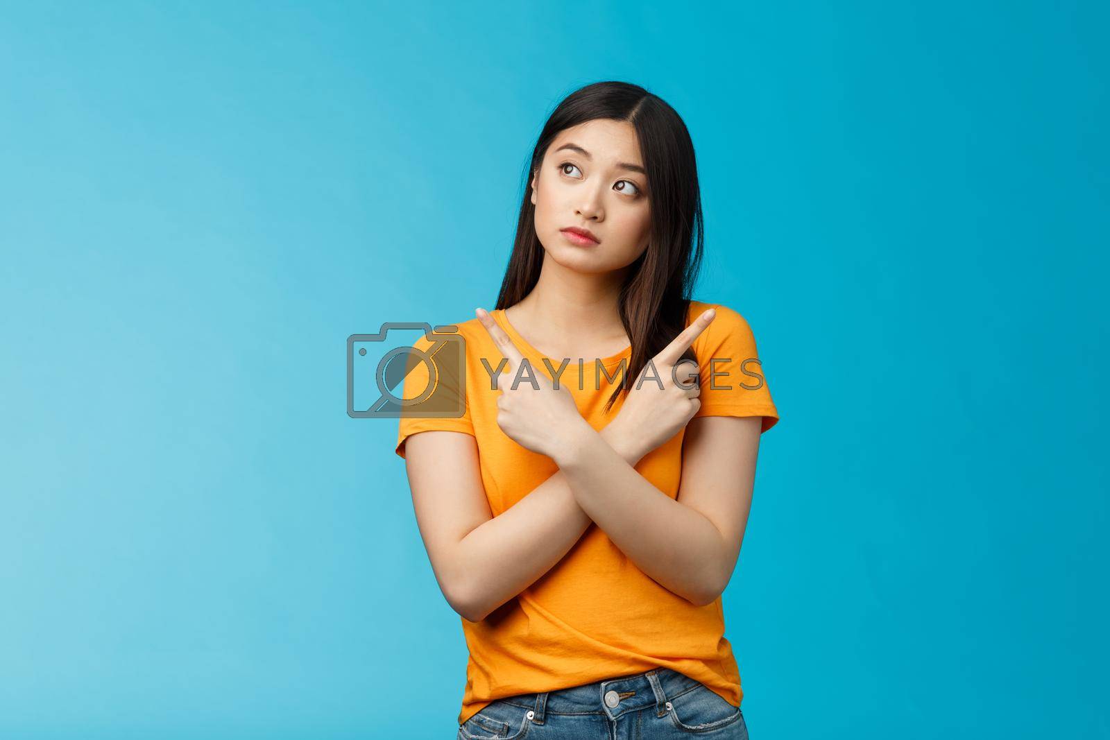 Royalty free image of Confused cute asian female student thinking what do, look sideways thoughtful, cross hands chest pointing left and right, making choice, thinking what pick, taking important decision blue background by Benzoix