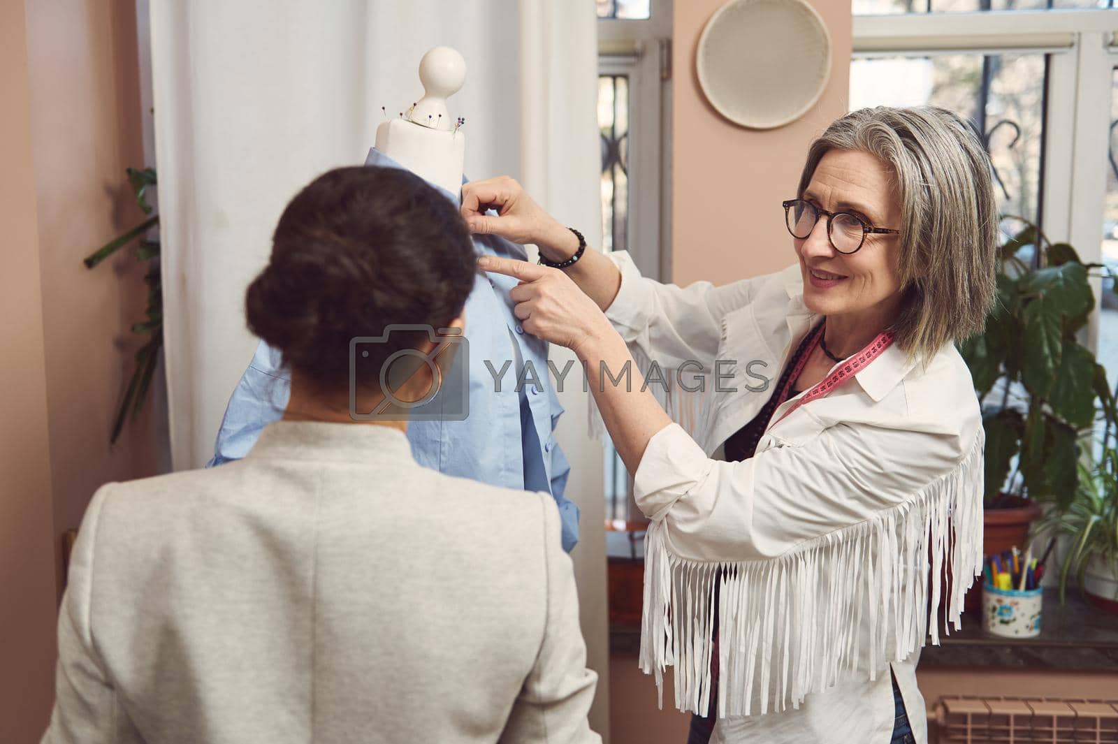 Royalty free image of Skilled team of fashion designers tailors try on the new collection of women's dress shirt on a mannequin and work to improve the style and design of the new garments in the tailoring atelier by artgf