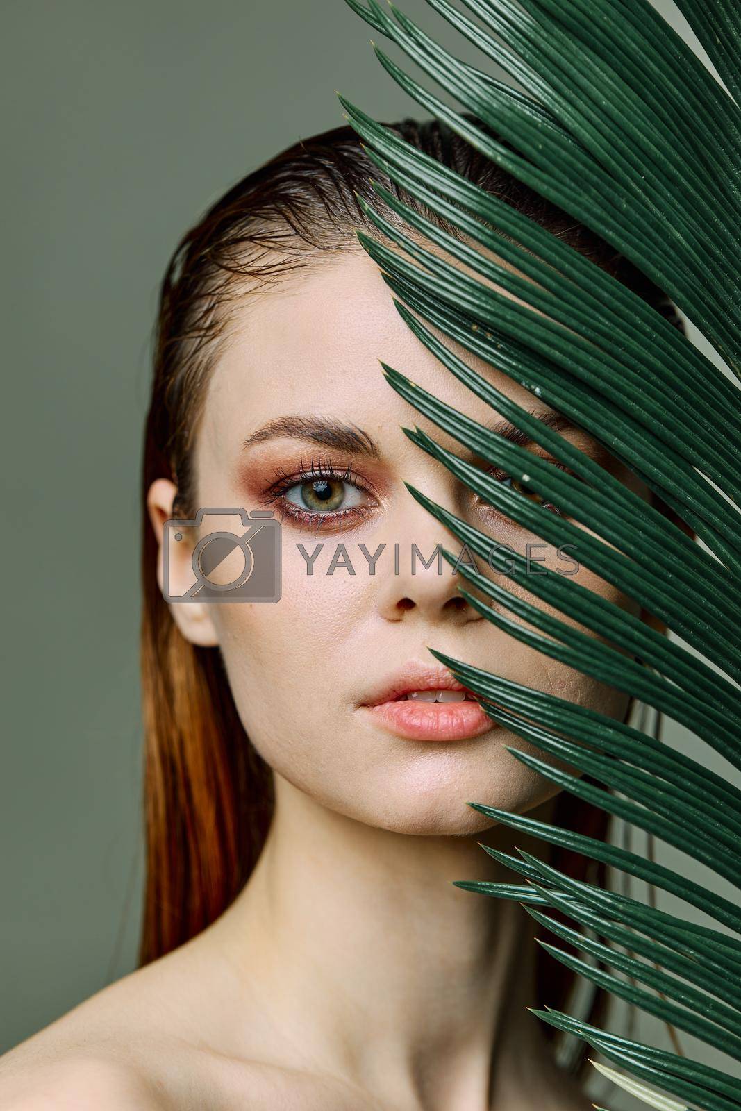 Royalty free image of a close beauty portrait of a beautiful woman standing holding a tropical palm leaf in her hand, bringing it to her face, looking into the camera. Vertical photo without retouching of problem skin by Vichizh