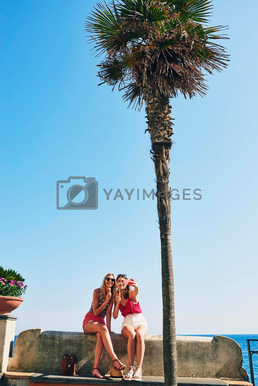 Royalty free image of Two friends taking selfies eating ice cream on beach using smart phone to connect on adventure travel by YuriArcurs