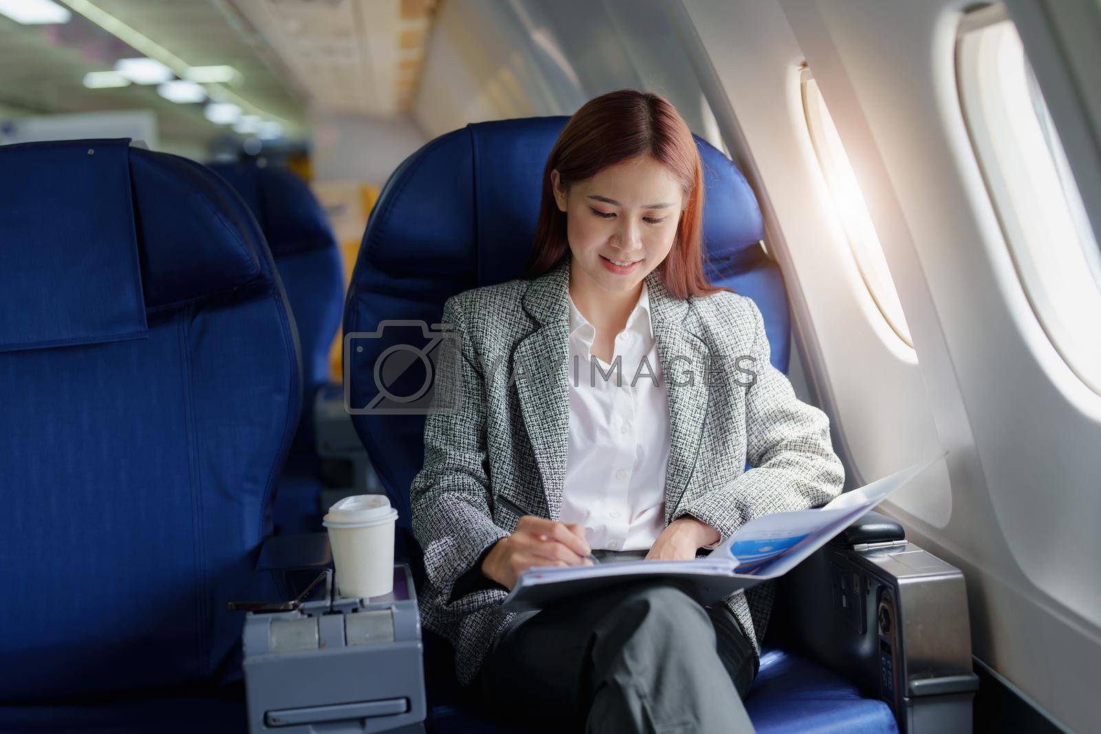 Royalty free image of portrait of A successful asian business woman or female entrepreneur in formal suit in a plane sits in a business class seat and uses a pen with documents for work during flight by Manastrong
