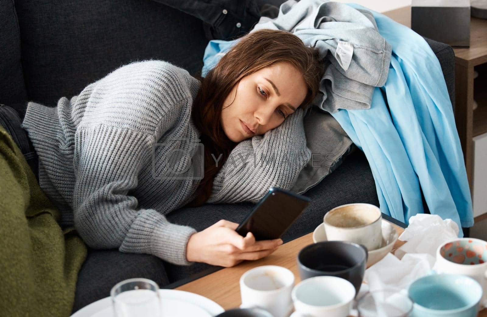 Royalty free image of When it borders obsession. Shot of a young woman lying on her messy couch looking depressed and using her smartphone. by YuriArcurs