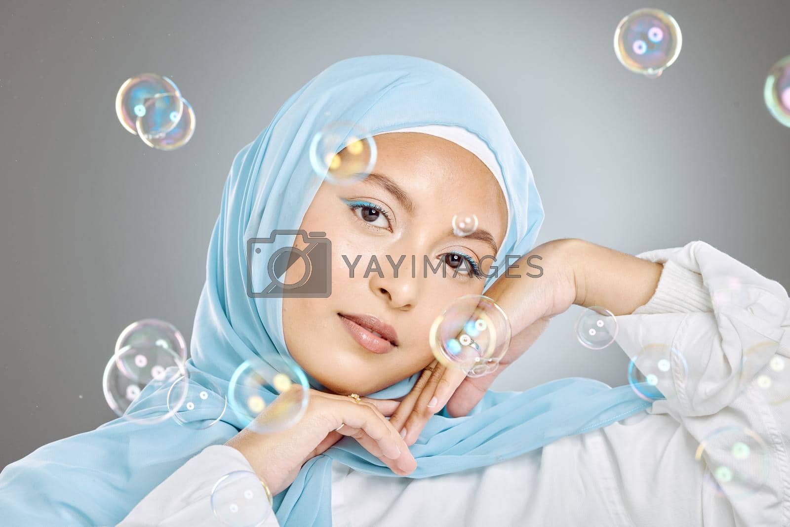 Royalty free image of Portrait of a beautiful muslim woman wearing blue hijab headscarf against grey background. Arab female in a happy daydream and fantasy surrounded by the pure delight and innocence of playful bubbles by YuriArcurs