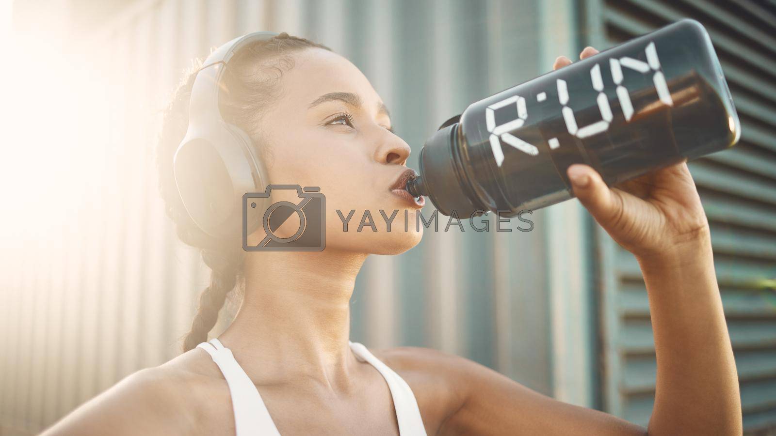 Royalty free image of One fit young hispanic woman wearing headphones and taking a rest break to drink water from a bottle while exercising outdoors. Female athlete quenching thirst and cooling down after run and training workout in an urban setting by YuriArcurs