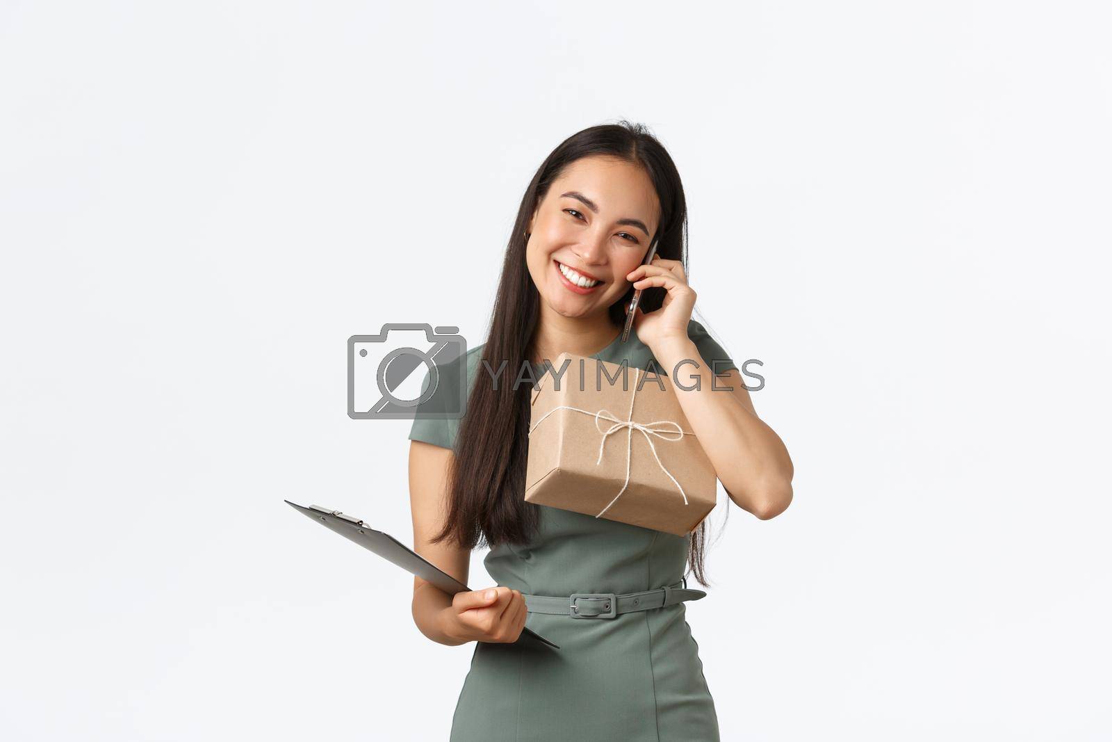 Royalty free image of Friendly smiling and polite asian female shop owner, managing small business, carry clipboard and package box for shipping, answering clients call, talking with customer, checking info for delivery by Benzoix