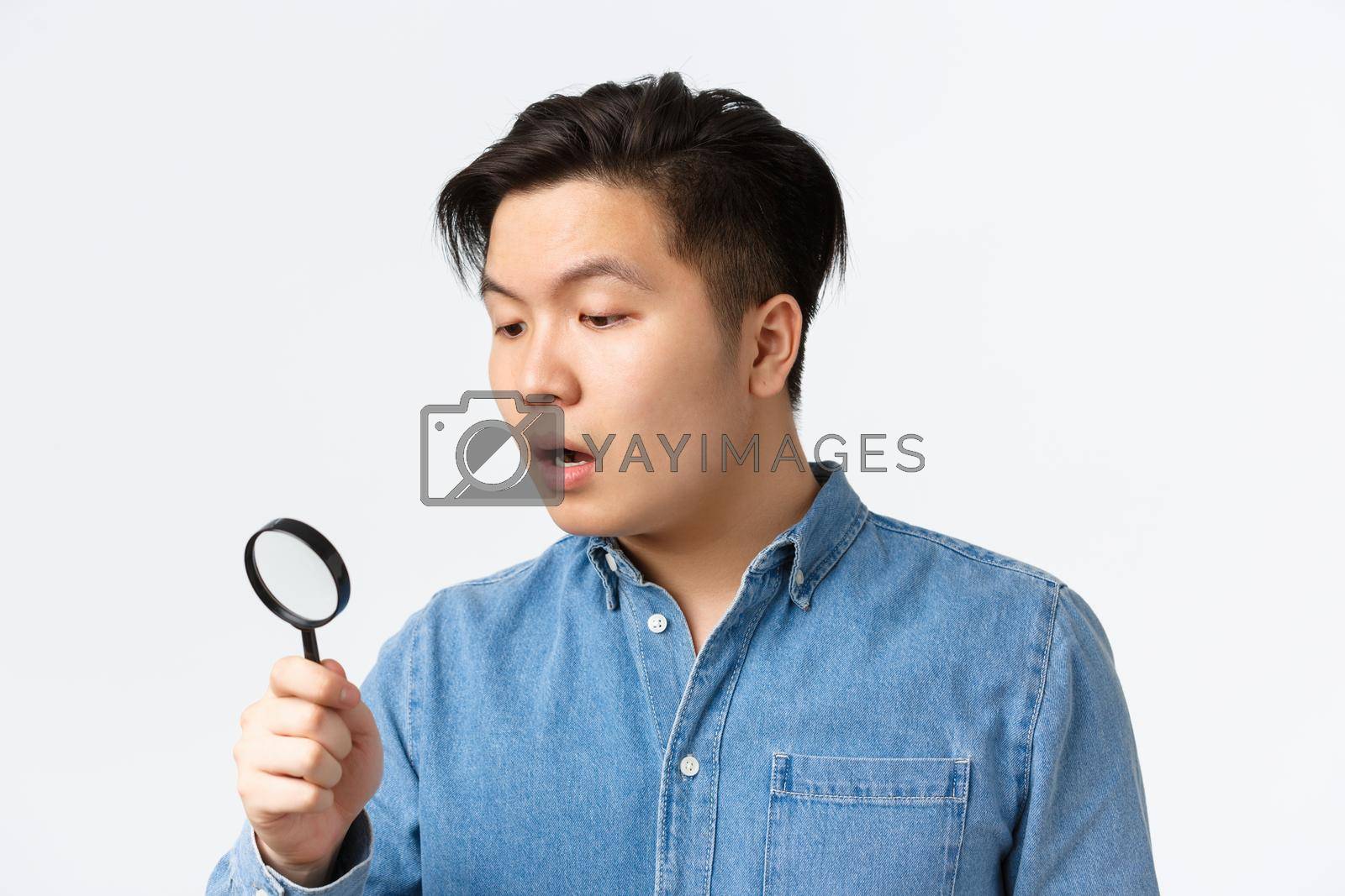 Royalty free image of Close-up of young asian guy looking curious and focused, searching for something, looking through magnifying glass down, open mouth intrigued, standing white background by Benzoix