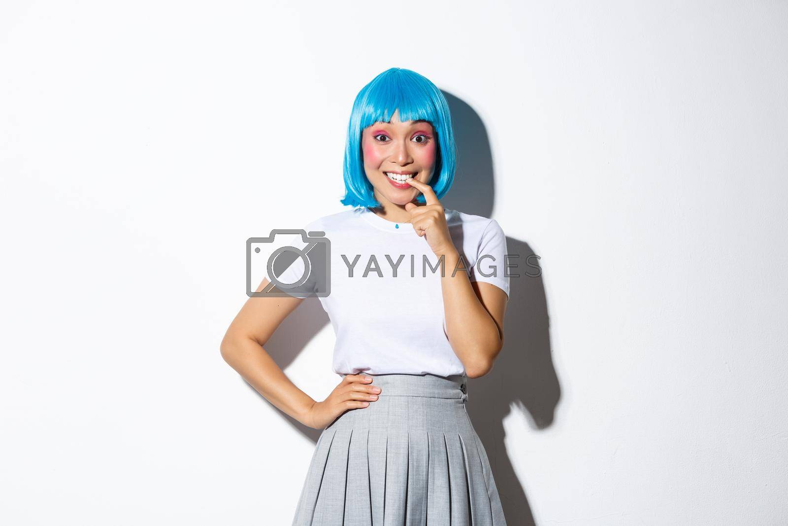 Royalty free image of Portrait of girl tempted to try something, looking with desire at camera, biting finger and smiling excited, wearing blue party wig for halloween, standing over white background by Benzoix