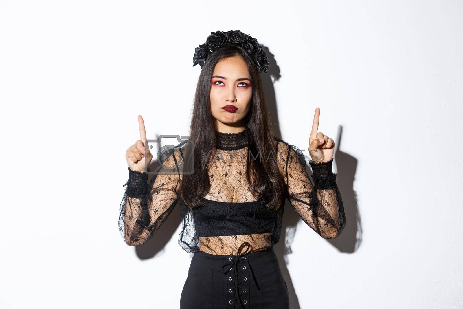 Royalty free image of Grumpy asian female in witch costume looking disappointed and upset, pointing fingers up and sulking displeased, standing over white background in halloween costume by Benzoix