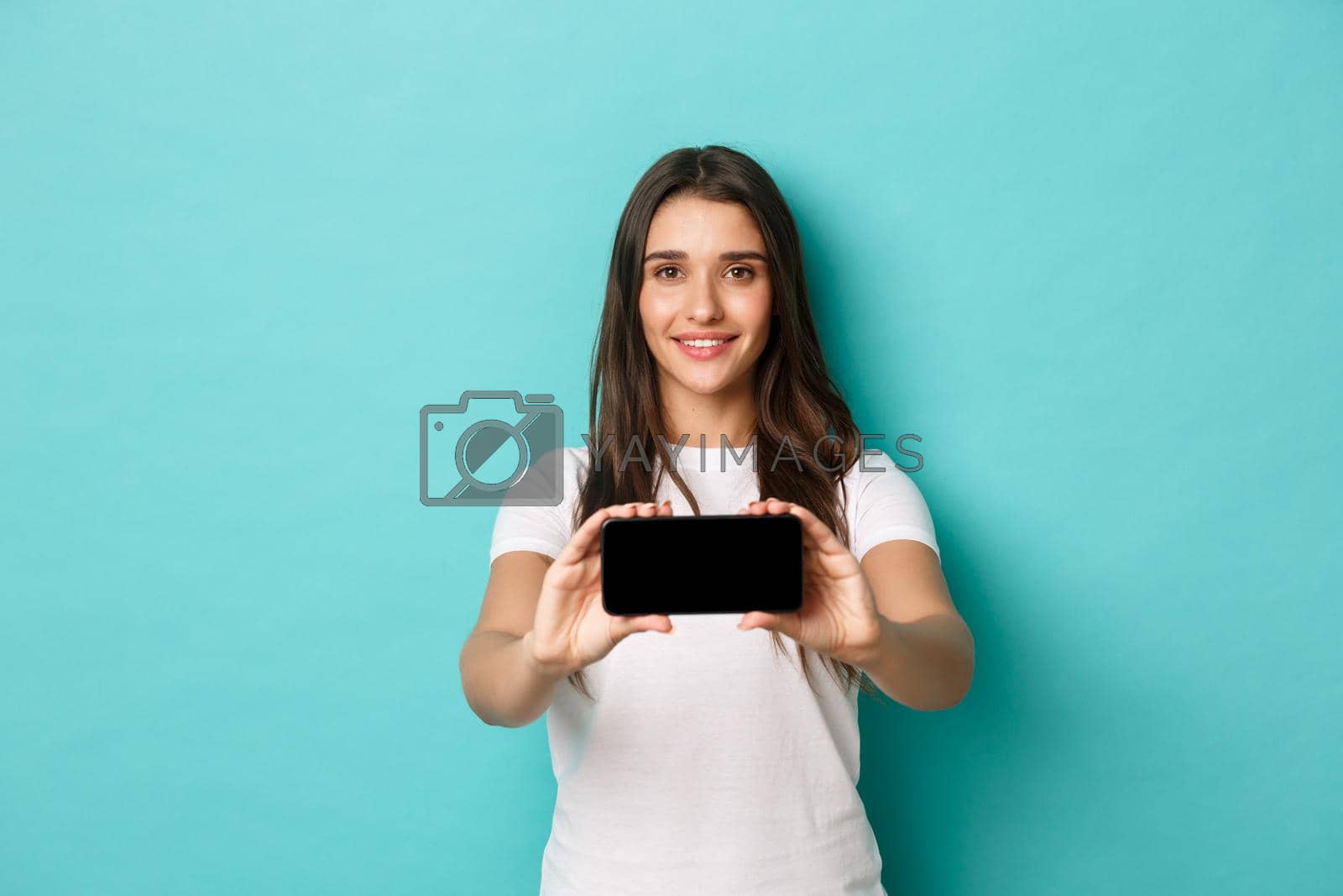 Royalty free image of Portrait of young rpetty woman in white t-shirt, showing smartphone screen horizontally, standing pleased over blue background by Benzoix