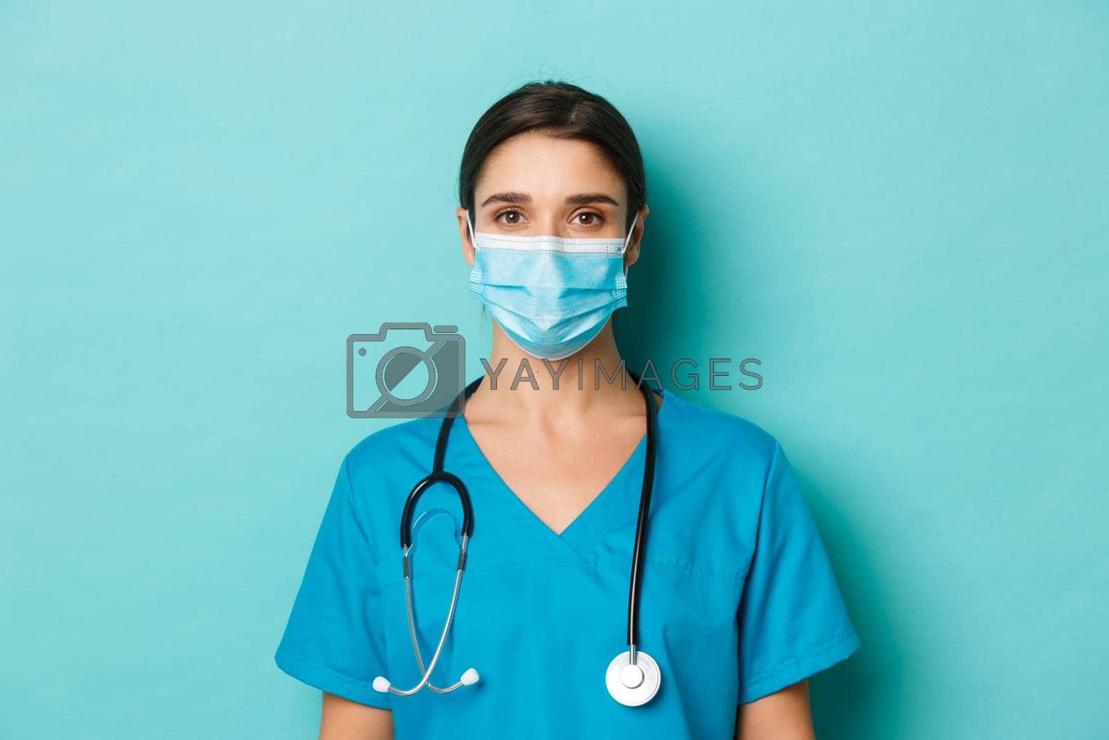 Royalty free image of Concept of covid-19 and quarantine concept. Close-up of young female doctor in scrubs and medical mask, looking at camera, standing over blue background by Benzoix